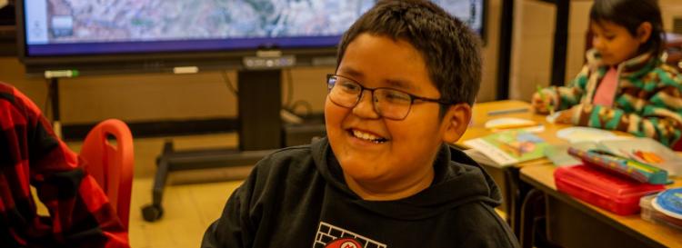 A smiling boy sits at a classroom table, looking happy and engaged in his learning environment.