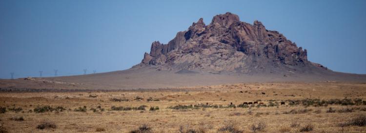 A large rocky mountain stands alone in the vast, dry desert landscape under a clear blue sky.