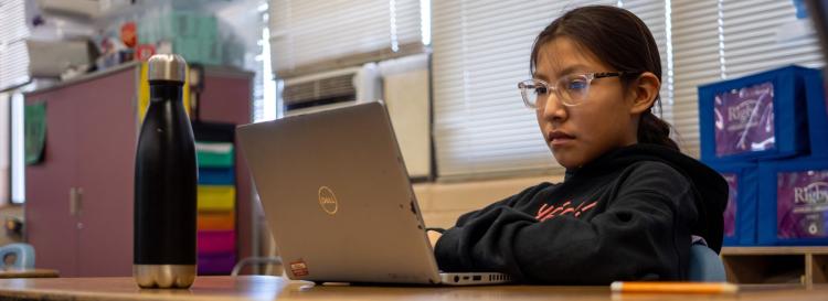 A student in glasses sits at their desk, intently working on a laptop, surrounded by books and school supplies.