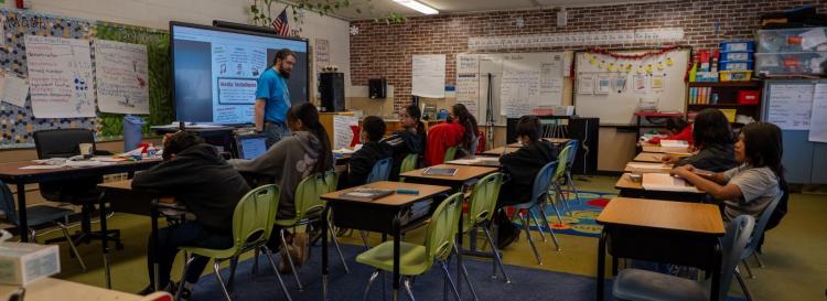 Students seated at desks in a classroom while a teacher stands at the front, leading the lesson.