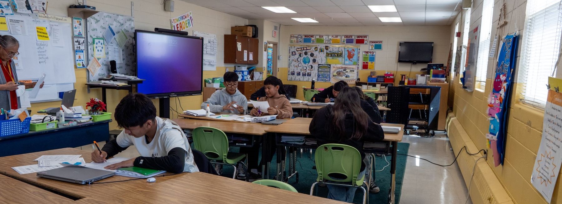 A classroom scene with students at desks and chairs, focused on their work and interacting with each other.