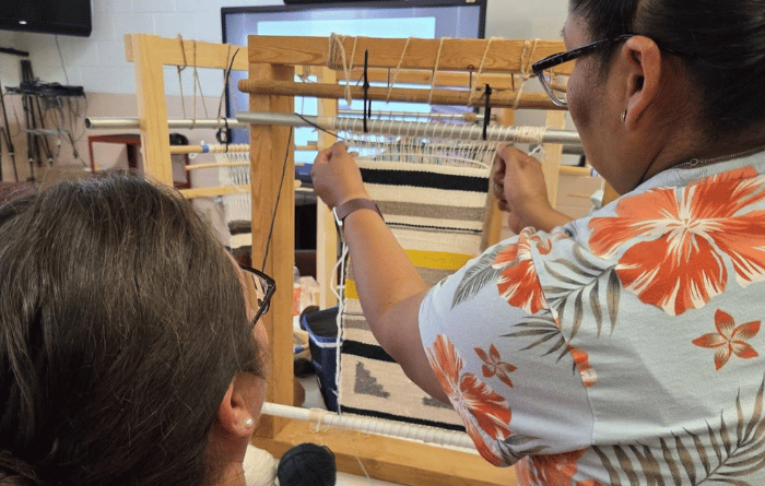 Two people work together at a floor loom during a weaving workshop, with one person threading or adjusting the warp while the other watches closely as a black-and-white textile takes shape.