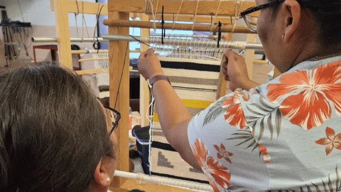 Two people work together at a floor loom during a weaving workshop, with one person threading or adjusting the warp while the other watches closely as a black-and-white textile takes shape.